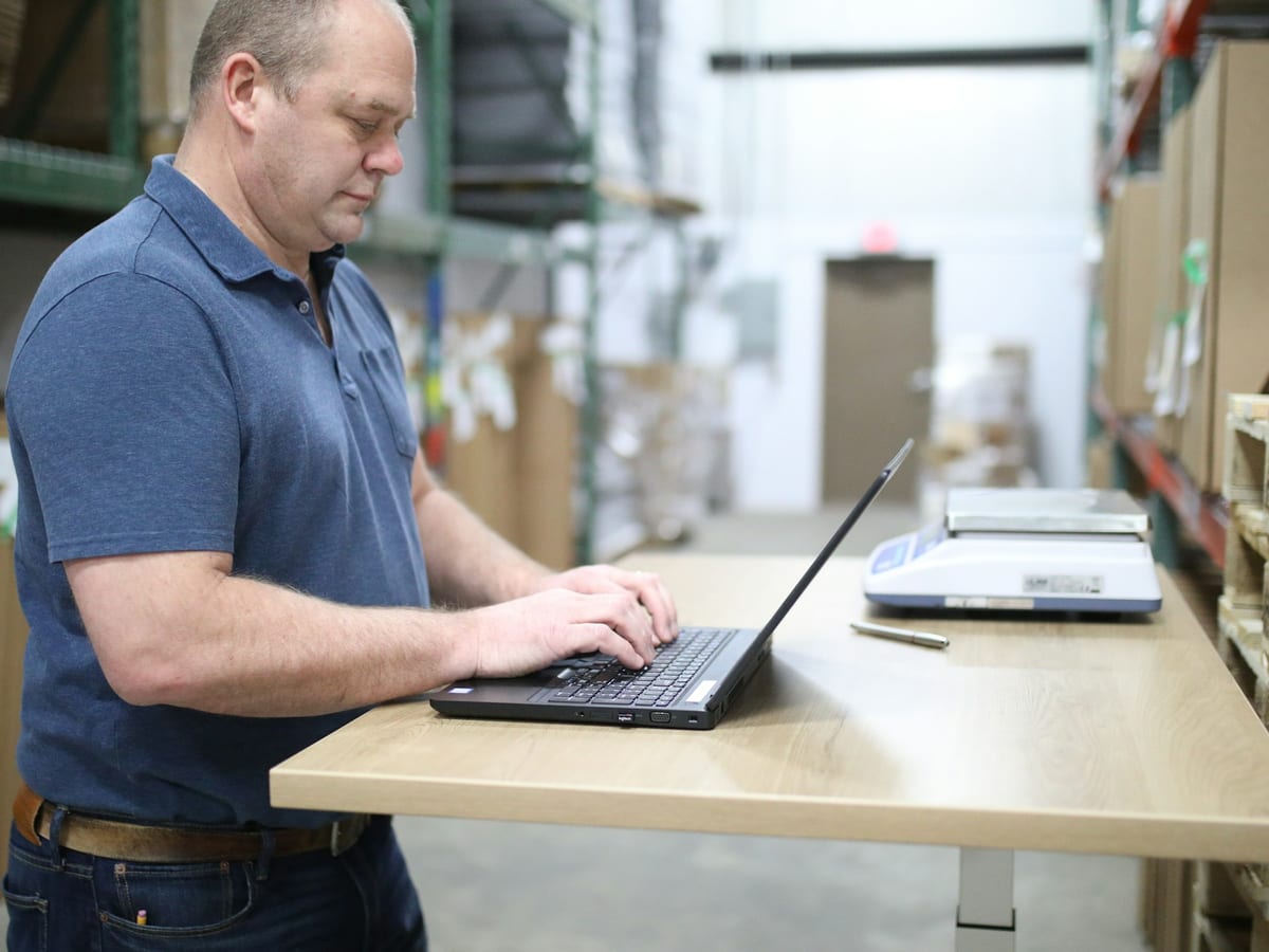 Materials manager working on a laptop at a standing desk in a stocked warehouse