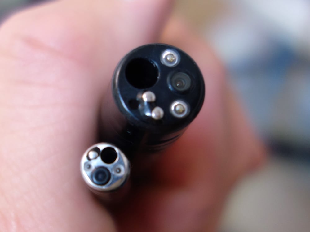 Close-up of two used endoscopic camera tips held between fingertips, showing the distal lenses, illumination ports, and a working channel with visible surface wear