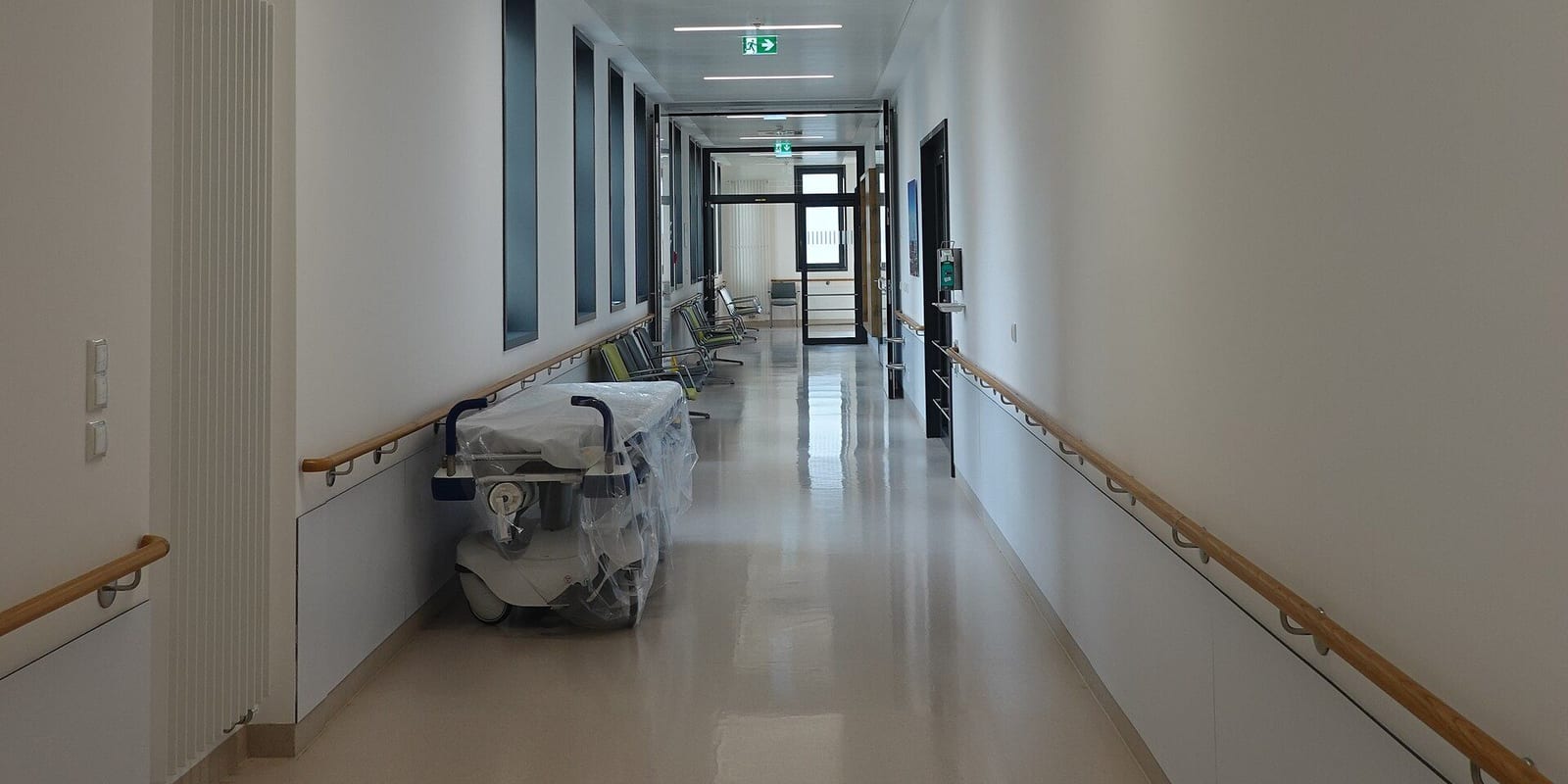 Hospital corridor with handrails, a plastic-wrapped stretcher and a row of empty seats by glass doors