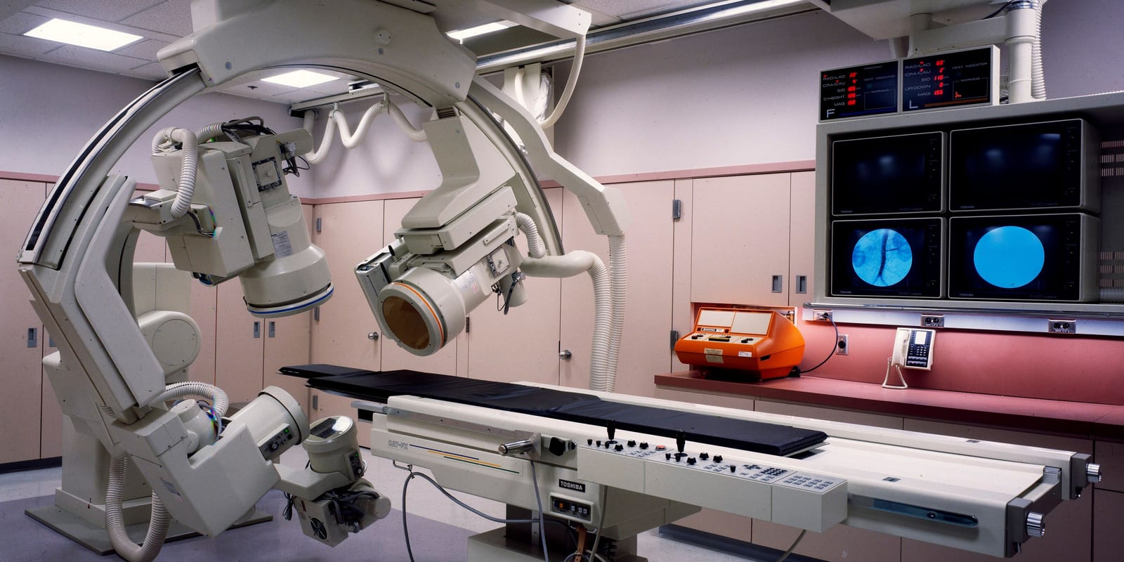 Cardiac cath lab: ceiling-mounted biplane X-ray arms over an empty table, wall monitors, cabinets