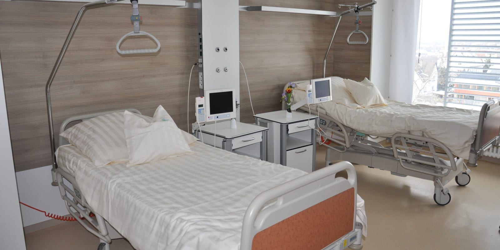 Two made hospital beds with overhead lifting poles, bedside monitors and a bright window
