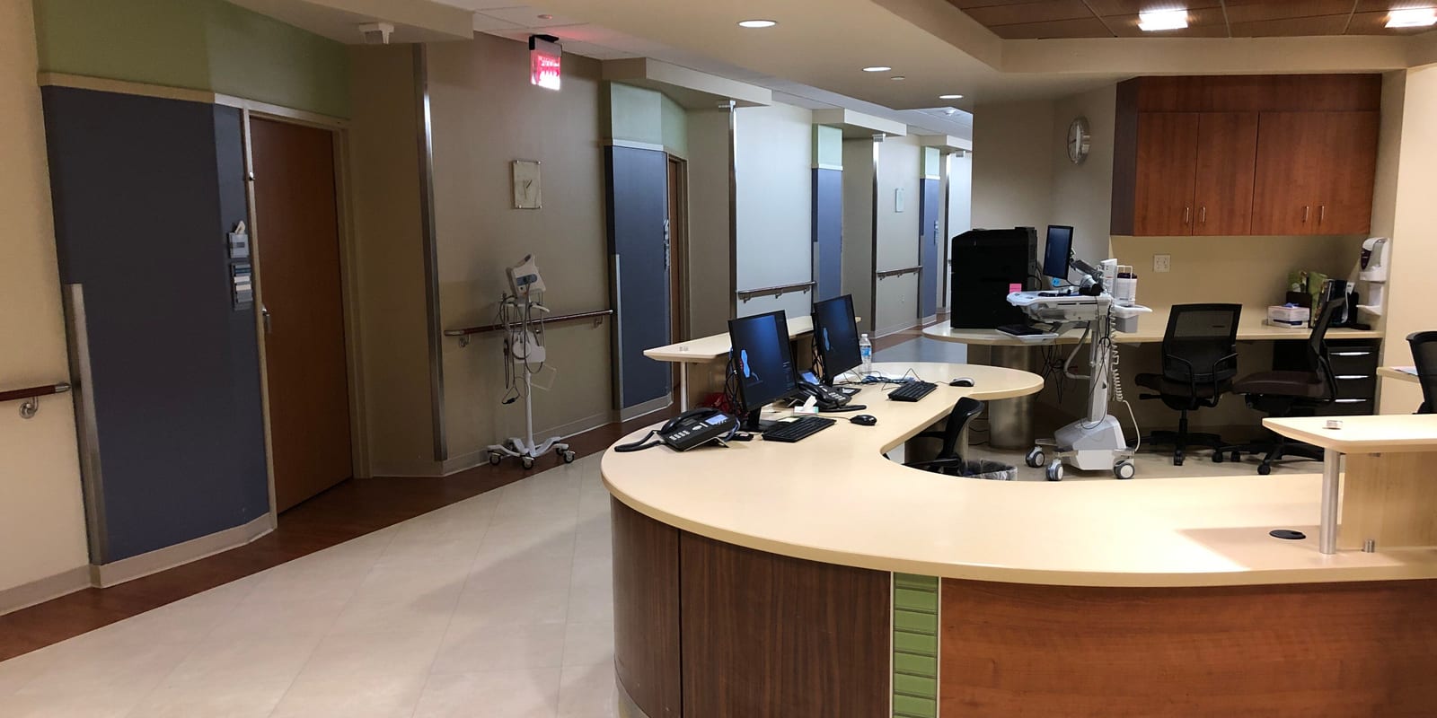 Empty curved nurse station with monitors and a computer cart facing patient-room doors