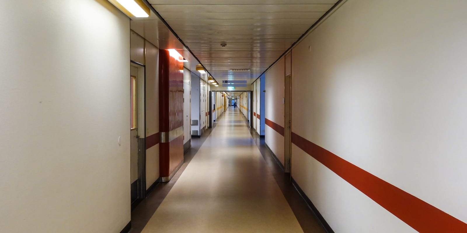 Long empty hospital corridor, ceiling lights, doors and a wall stripe receding to a vanishing point