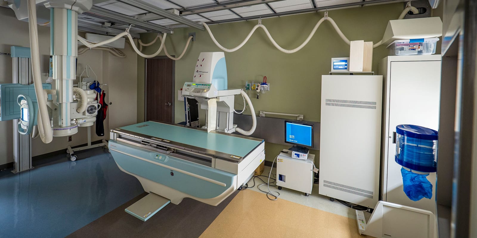 X-ray room: ceiling tube on rails, radiography table, wall stand, cabinets and control screen