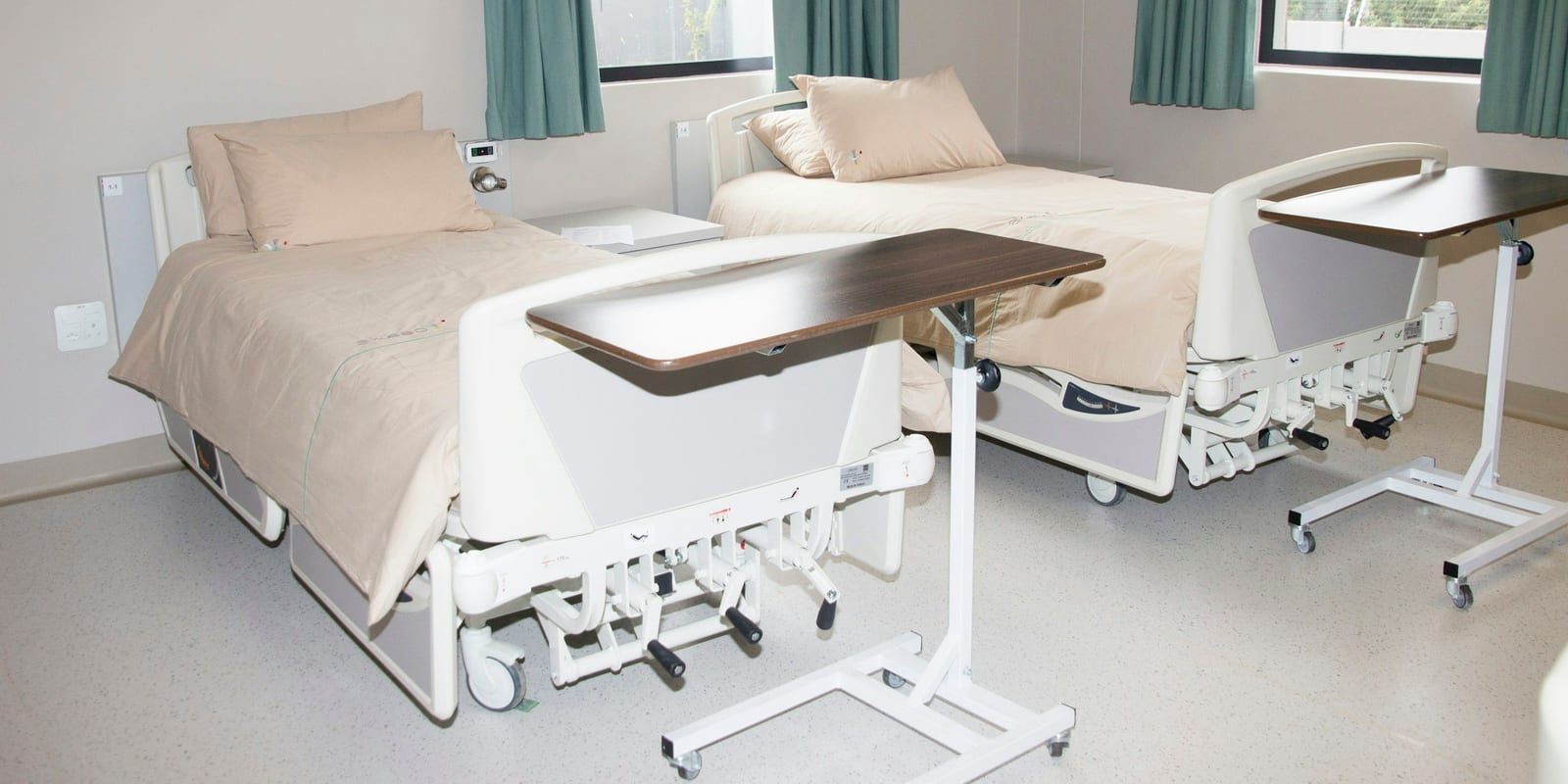 Two made hospital beds with overbed tables beside windows with green curtains