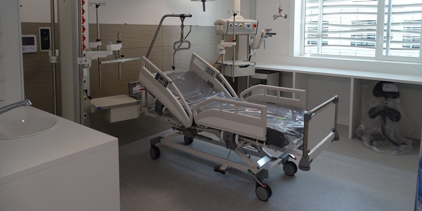 ICU room with a new bed still in plastic wrap, ceiling supply column, sink and window