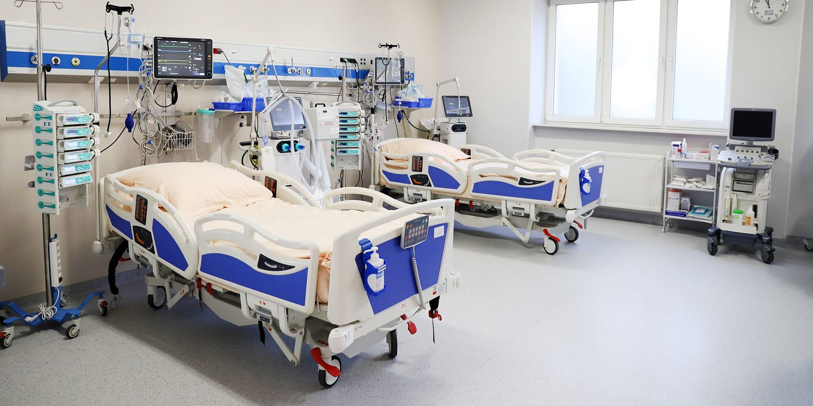 ICU bay: two empty beds, ventilator, infusion pump stacks, monitors on booms, ultrasound cart