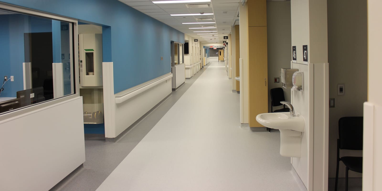 Emergency-department corridor with glazed bays, handrails, a wall sink and empty chairs
