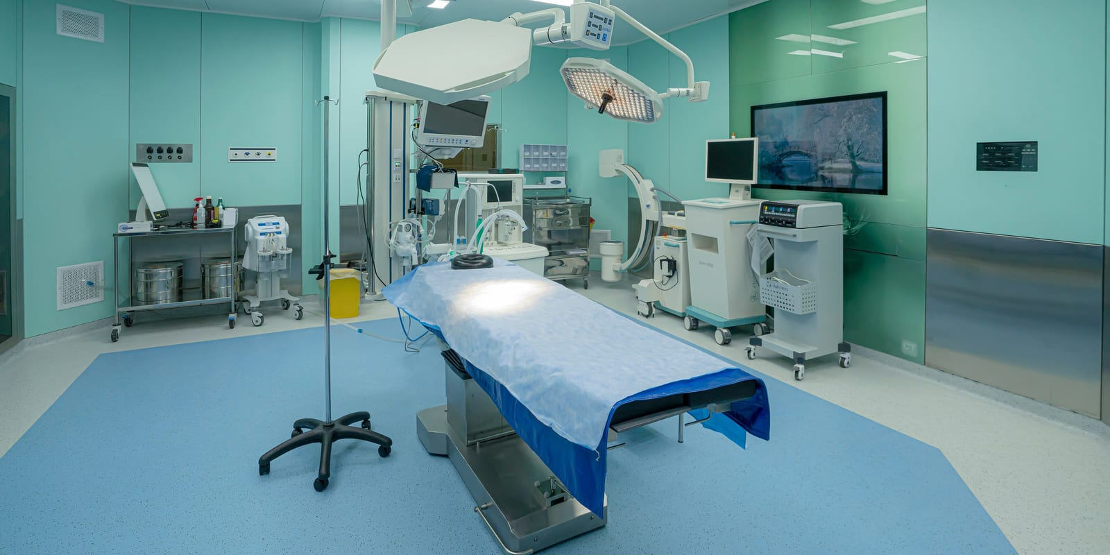 Empty operating room: draped operating table, twin LED surgical lights, C-arm and trolleys, wall screen