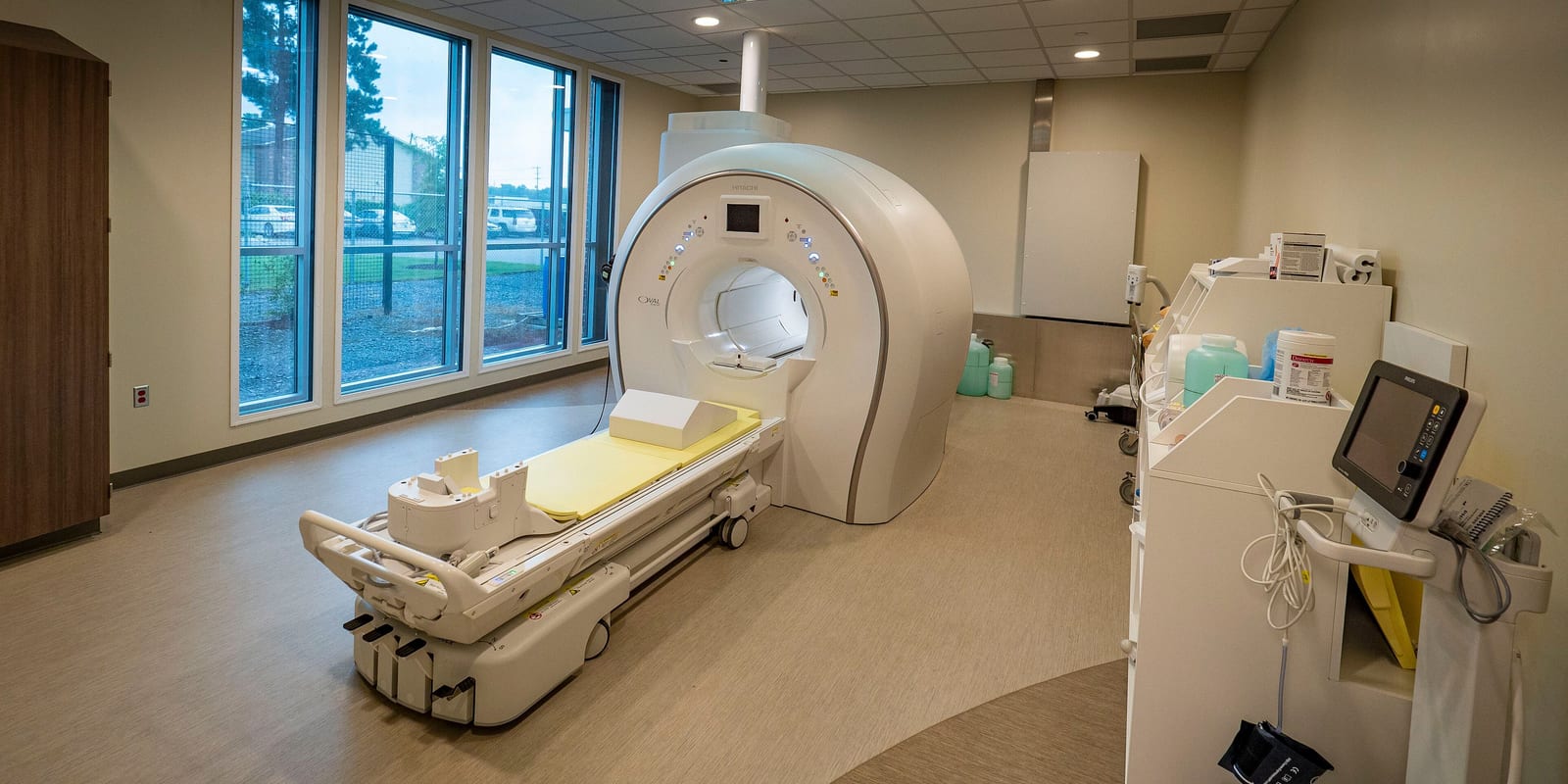 MRI room with windows: scanner and table, contrast injector cart and monitor beside it