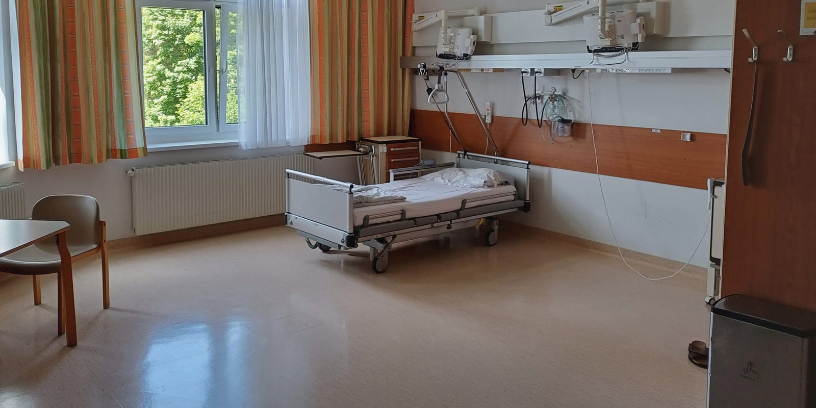 Bright patient room with an empty bed at the headwall rail, a table and chair by the window