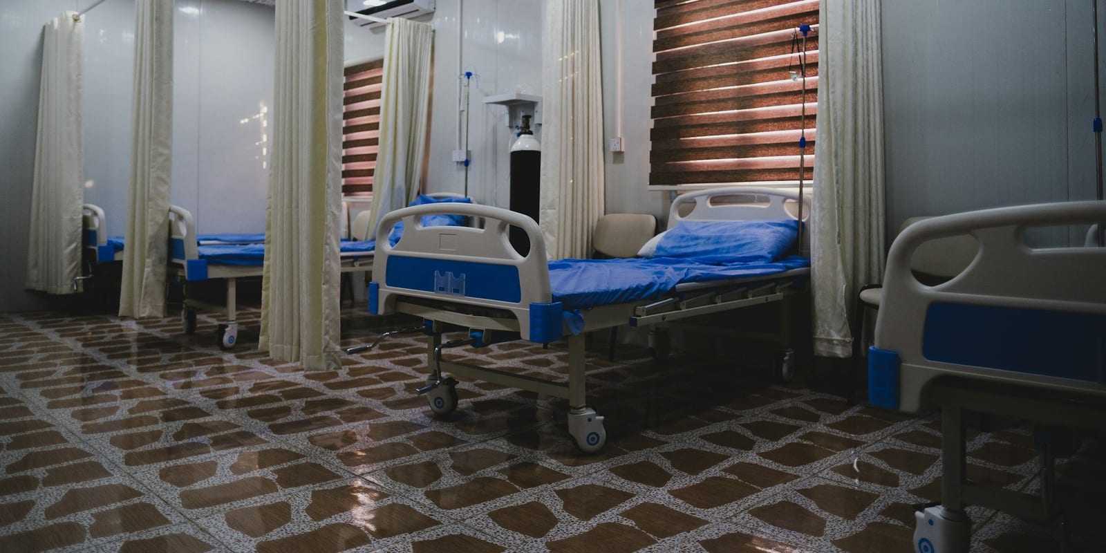 Ward of empty beds with blue mattresses separated by privacy curtains, window blinds behind