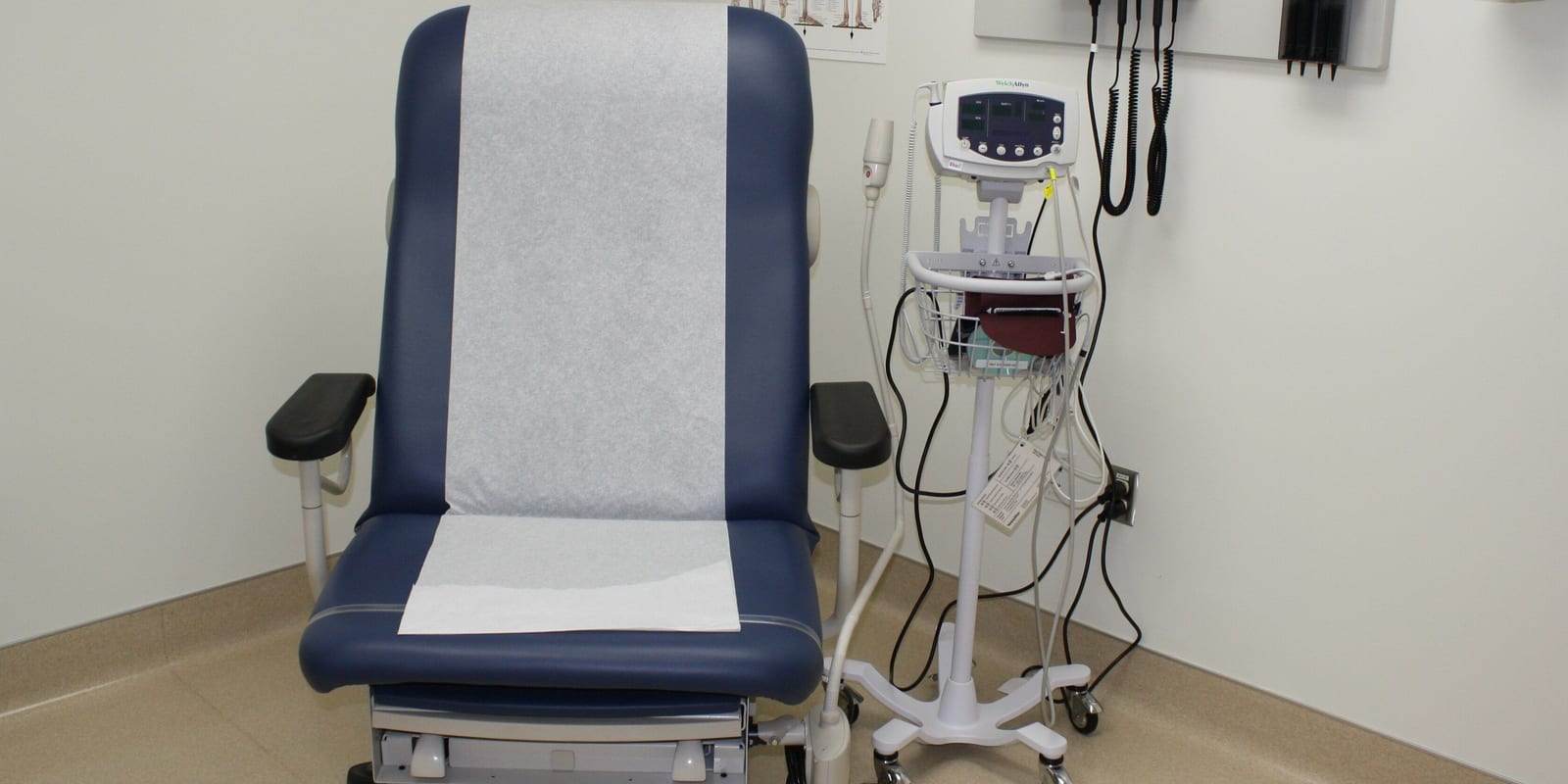 Examination room: paper-covered exam chair, wall diagnostic set, vitals monitor on rolling stand