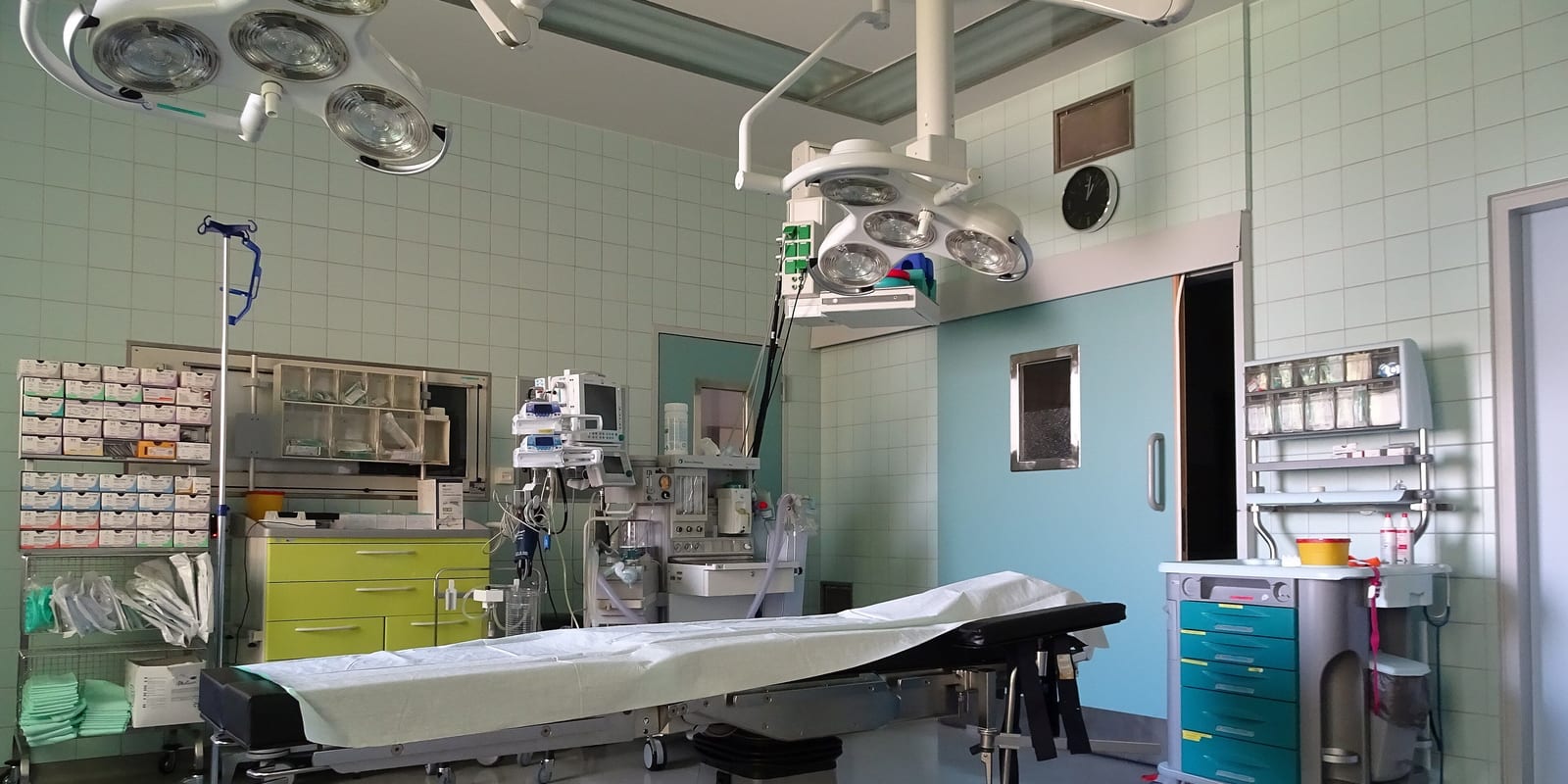 Operating room: draped table, two ceiling surgical lights, anaesthesia unit, tiled walls, supply shelving