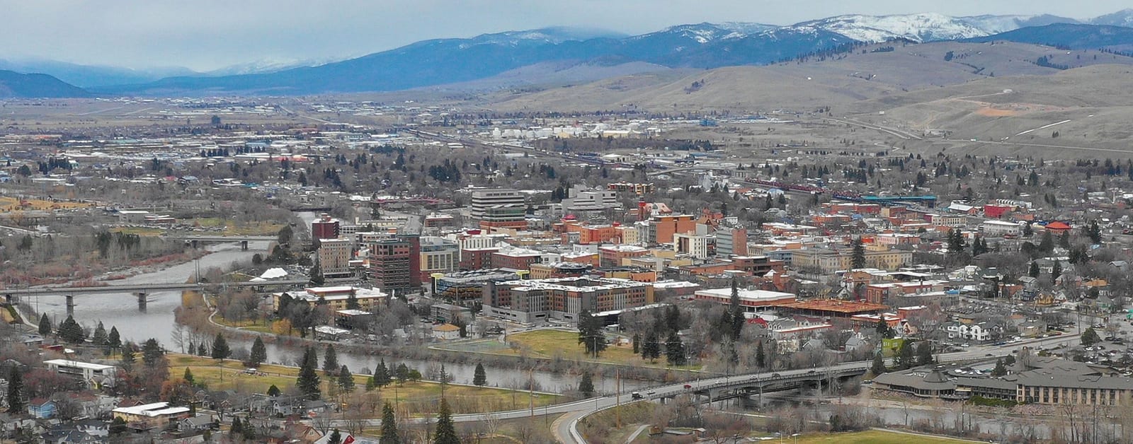 Aerial view of downtown Missoula beside the Clark Fork River, with bridges and snow-capped mountains beyond