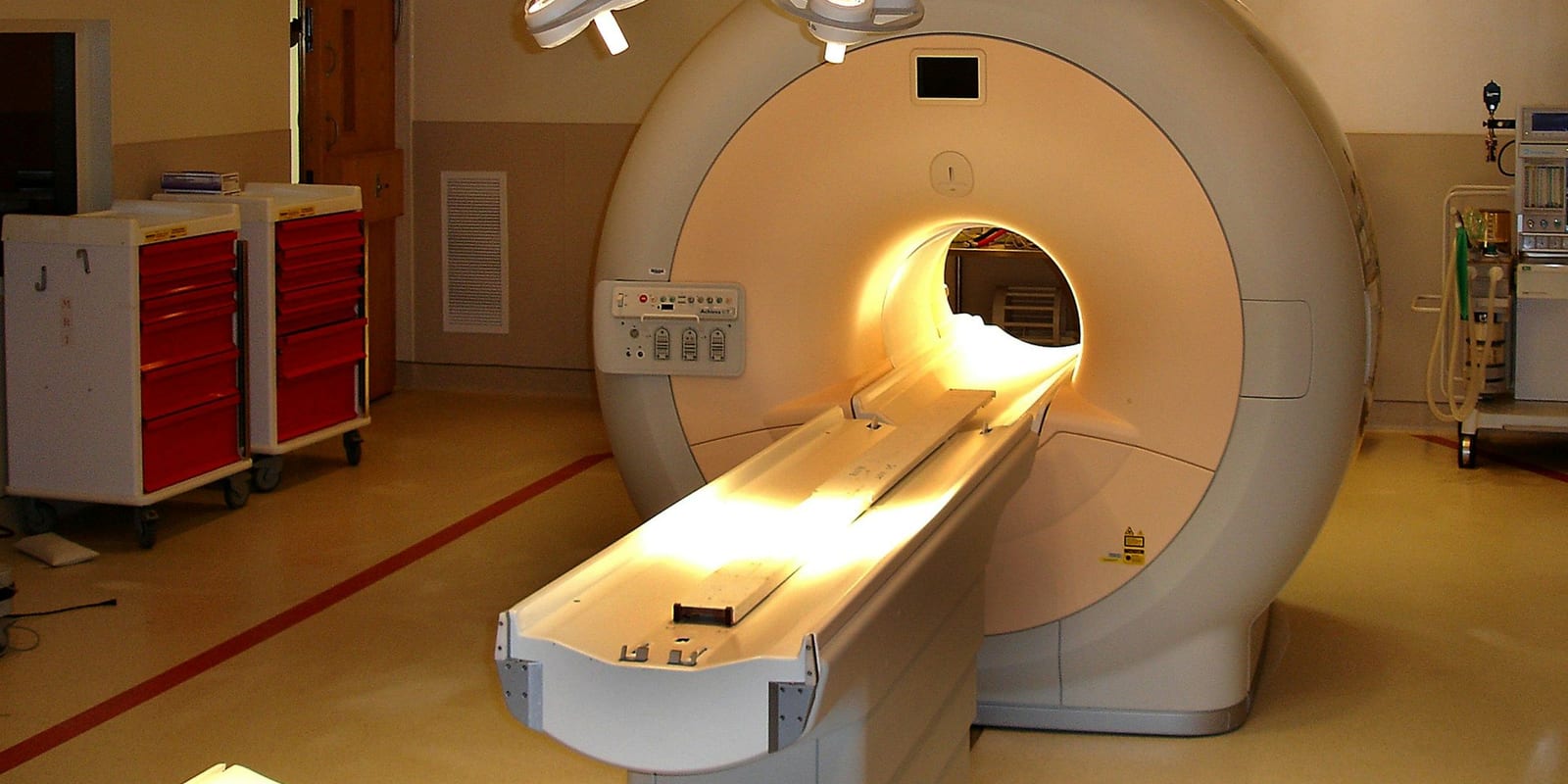 MRI scanner with lit bore and table, red supply carts and an anaesthesia machine at the sides