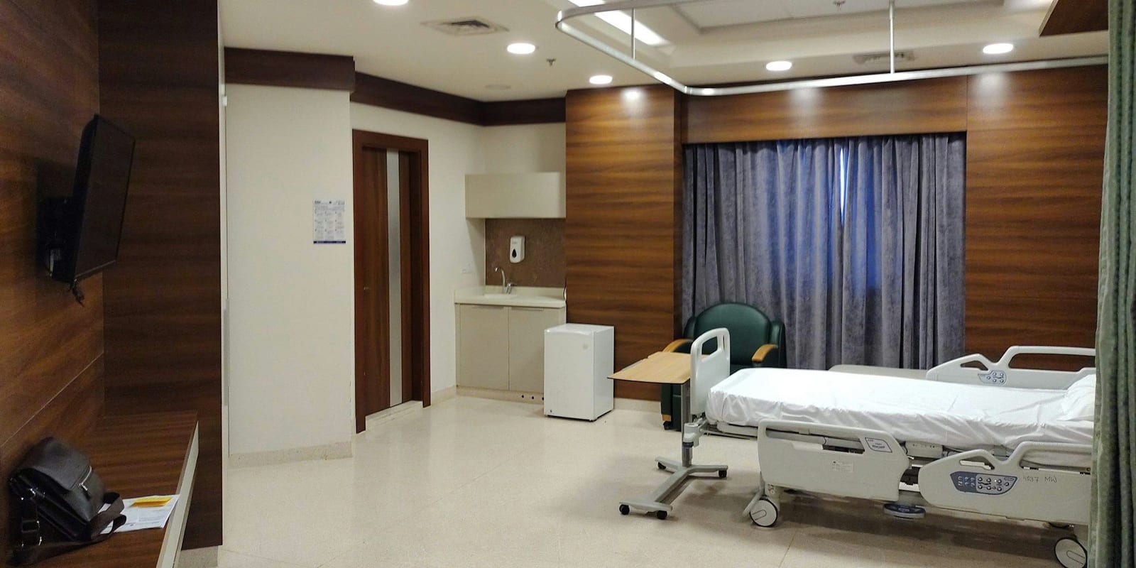 Private hospital room with a made bed, overbed table, armchair, sink and curtained window