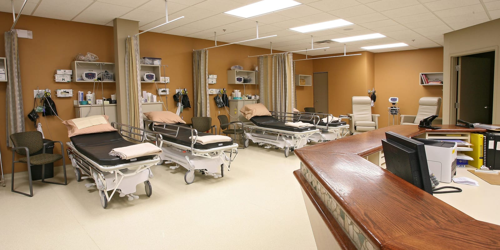 Empty post-anaesthesia recovery bay: row of stretchers, vitals monitors, curtain tracks, nurse desk