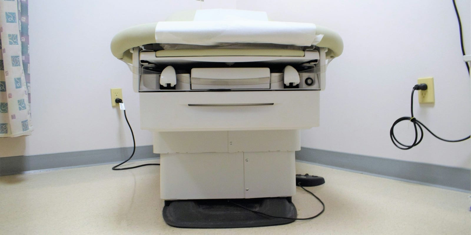 Motorised exam table with paper roll against a plain wall, patterned curtain and power cord