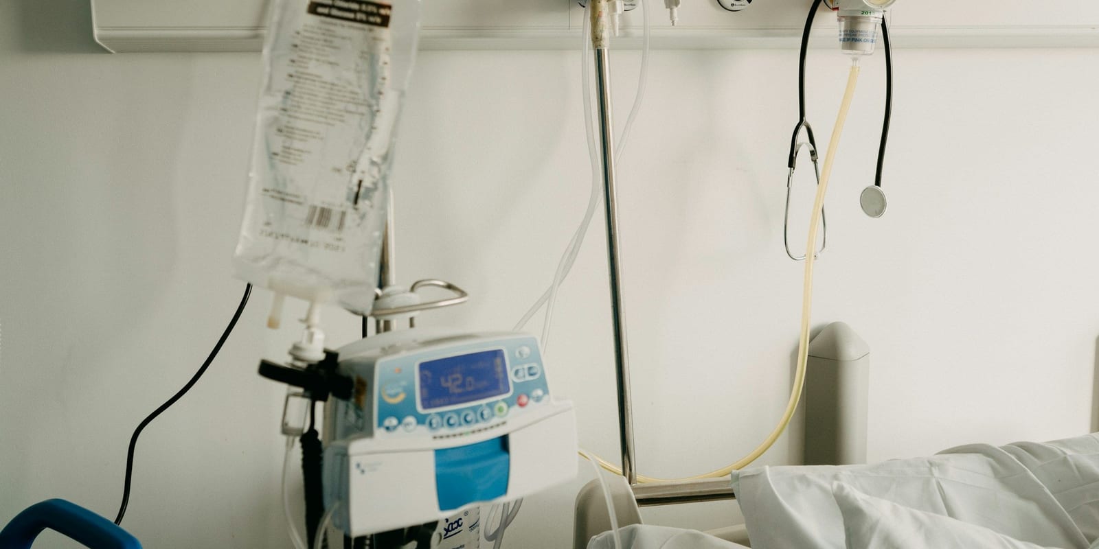 Infusion pump on an IV pole with fluid bag, wall stethoscope and gas outlet above bedding