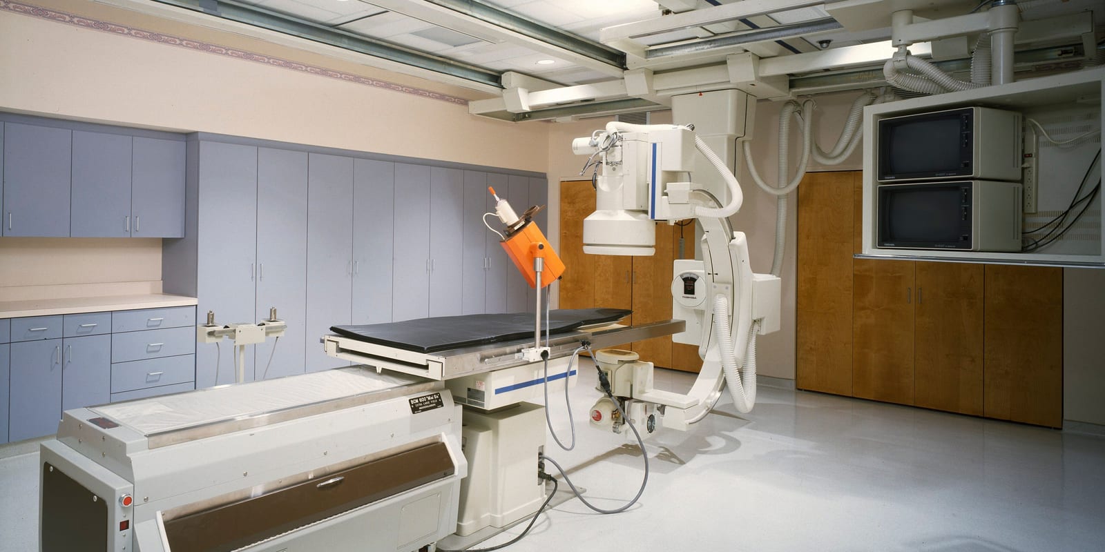 Fluoroscopy room: C-arm and ceiling tube over an empty table, cabinets and wall monitors