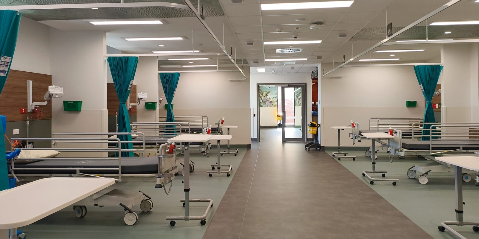 Open multi-bed ward bay: empty beds, over-bed tables, privacy curtains, aisle to a glazed door