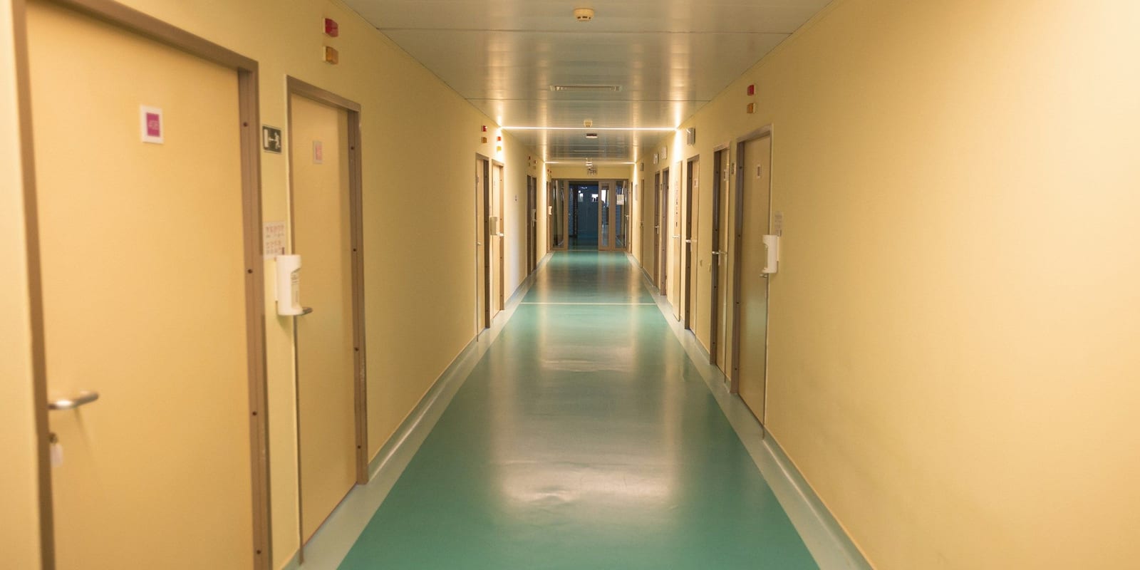 Long hospital corridor with closed room doors, call lights, sanitiser dispensers and a teal floor