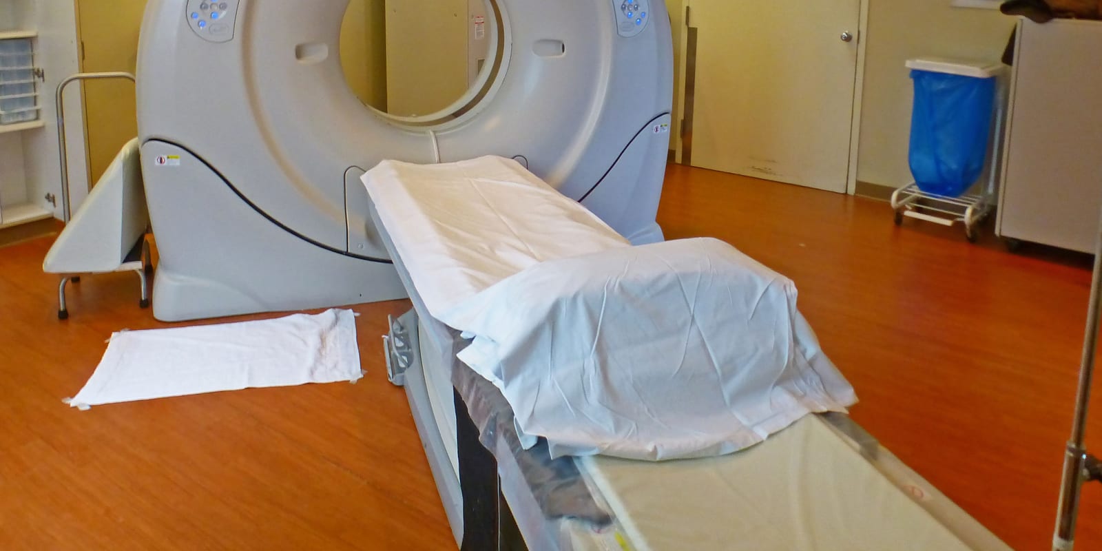 CT scanner with sheet-covered table, bin on a stand, cupboards and a closed door