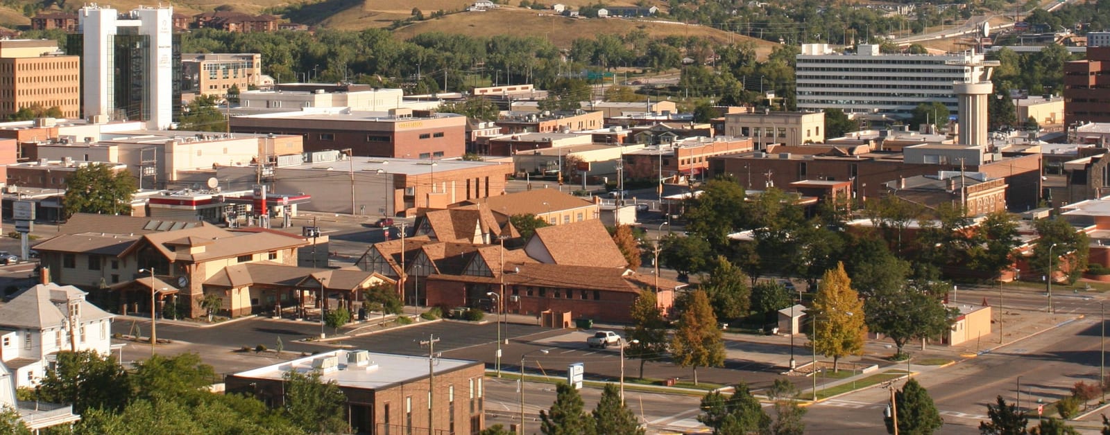 Downtown Rapid City rooftops and office buildings in warm low sunlight, with grassy foothills behind