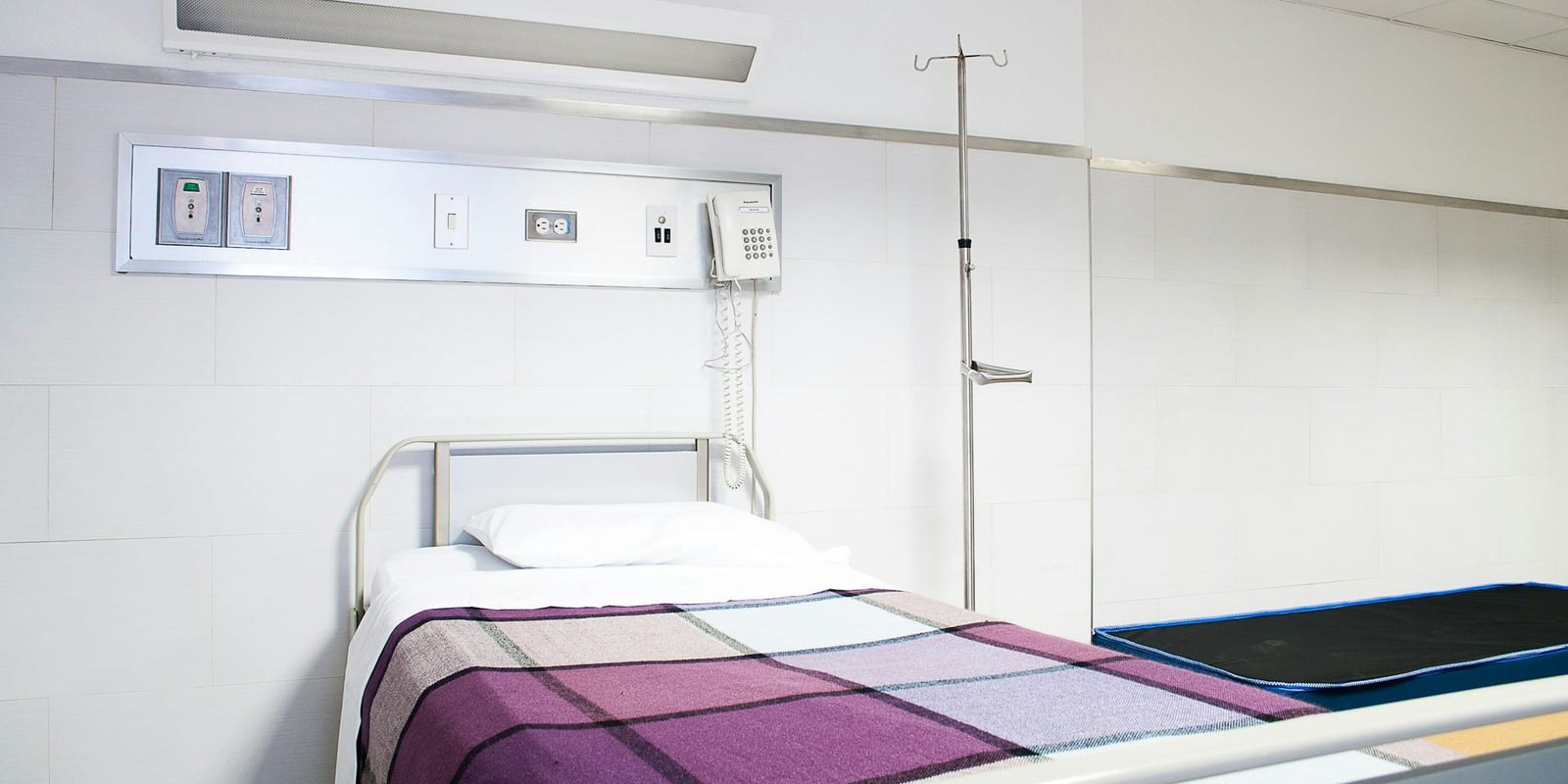 Empty hospital bed with a checked blanket under a headwall panel with outlets, phone and IV pole