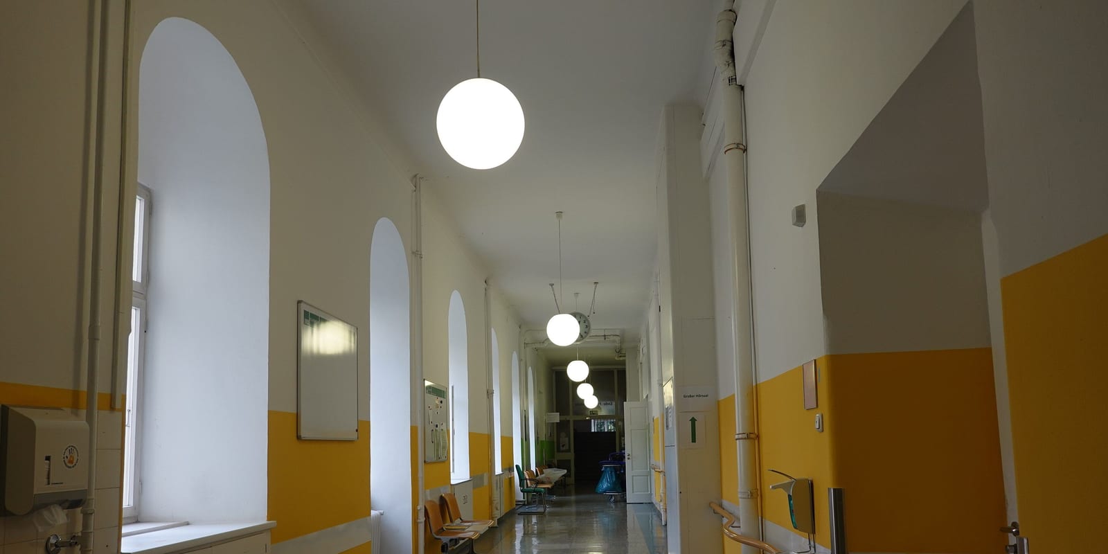 Vaulted hospital corridor with arched windows, globe lamps, handrails and empty benches