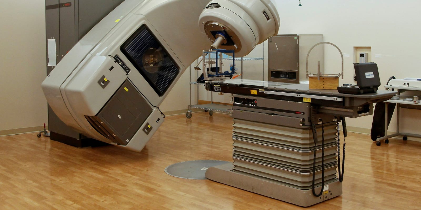 Linear accelerator gantry tilted over an empty treatment couch on a wood floor, carts and cabinets