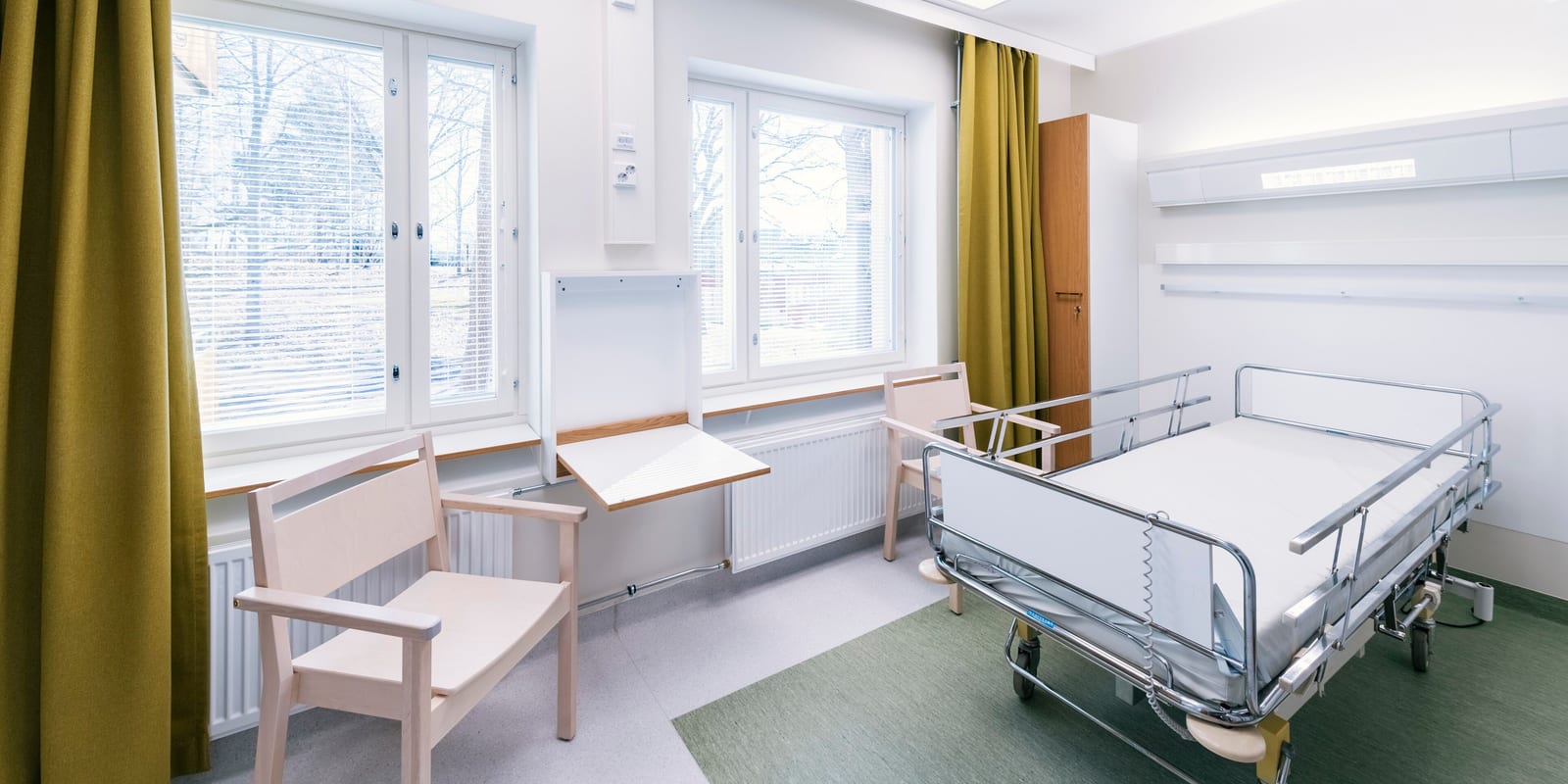 Bright, empty patient room: made hospital bed, wooden chairs, fold-down table, tall windows