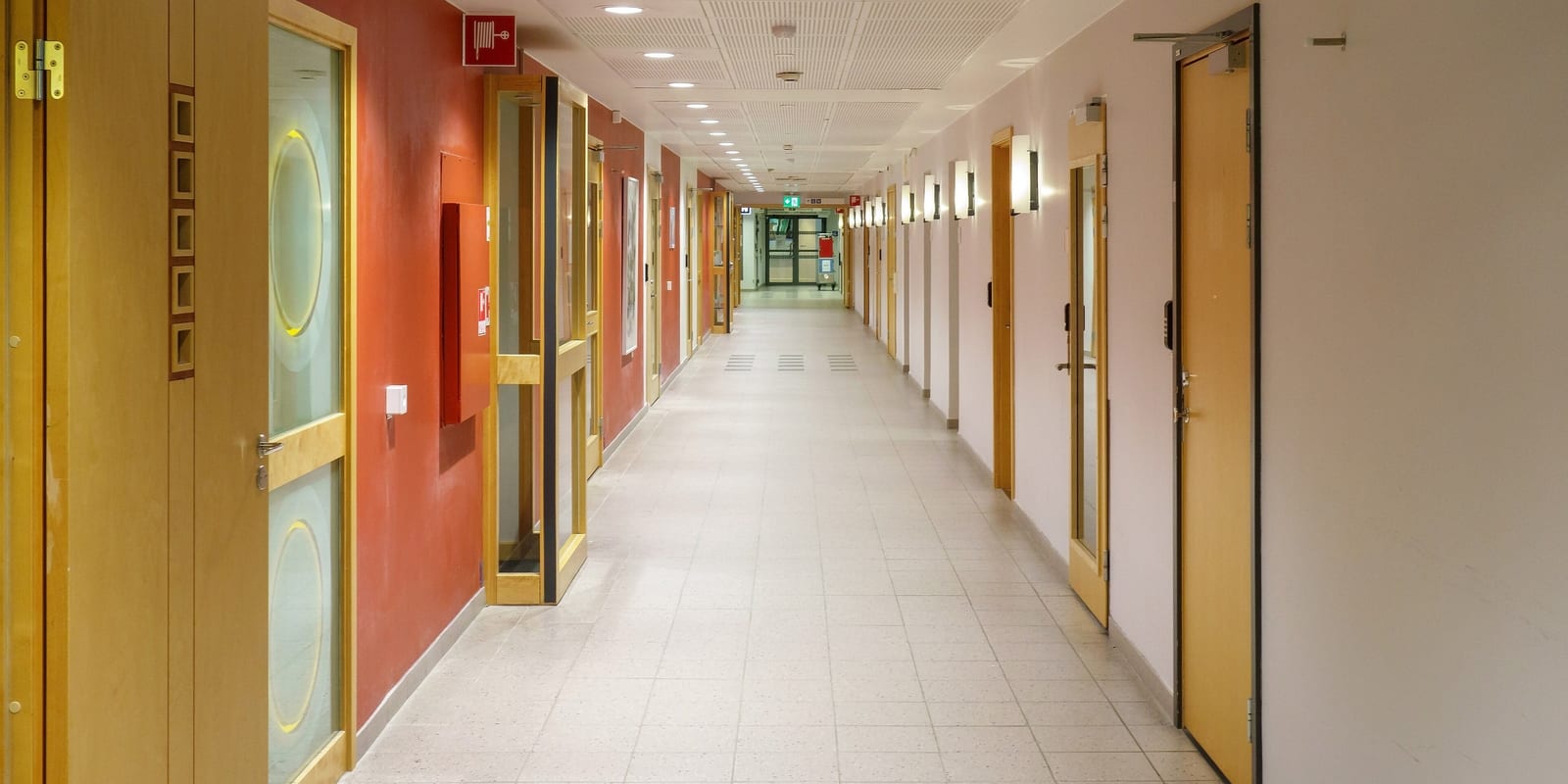 Hospital corridor lined with wooden doors and wall lights, tiled floor receding to glazed doors