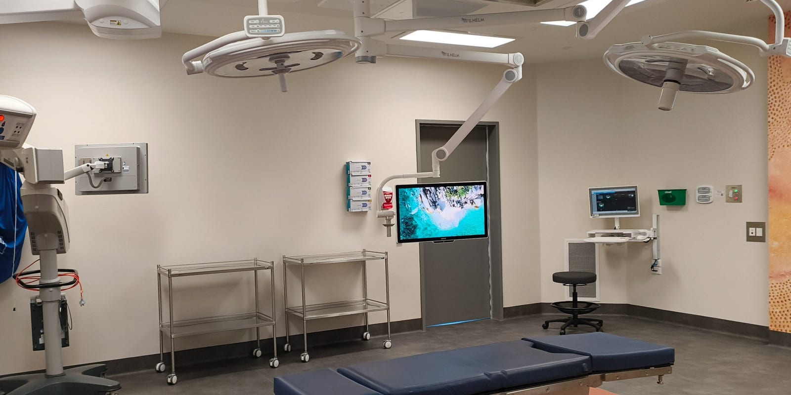 Empty operating room: two surgical lights, instrument trolleys, wall monitors, padded table in front