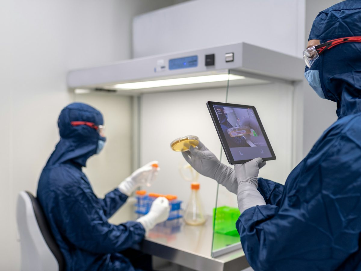 Technician working inside a laminar flow hood while a gowned colleague checks a tablet